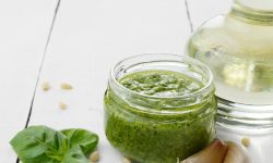 Basil pesto in a glass jar on white surface with basil leaves, pine nuts, garlic and olive oil in background